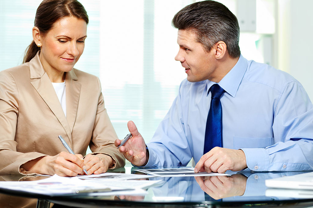 Мужчина и женщина сидят за столом, женщина ставит подпись на документах